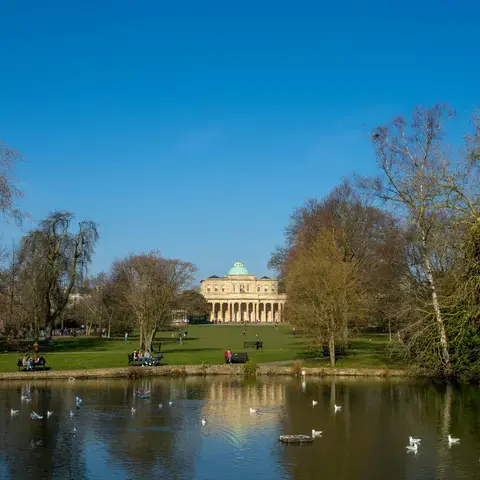 Reflections in a calm park lake surrounded by leafless trees, with a neoclassical building under a vivid blue sky