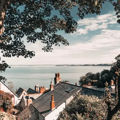 Seaside village with slate-roofed houses framed by trees, overlooking a calm sea under a partly cloudy sky