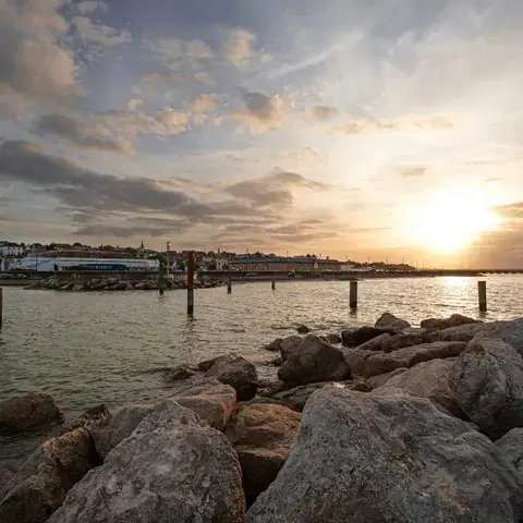 Sunset over a calm coast, waves hit the rocks gently. A serene moment by the sea as the town glows under the fading light