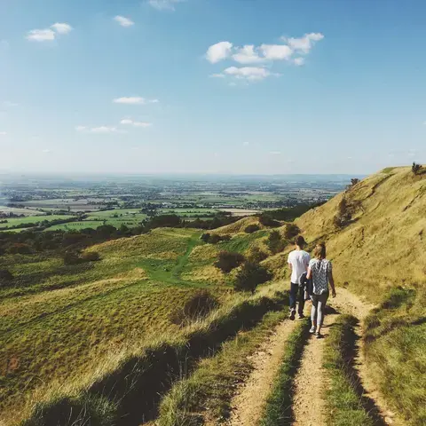 Two hikers walk a hillside trail overlooking green fields and a blue sky, surrounded by breathtaking natural scenery