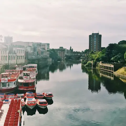Colorful boats moored on a tranquil river with historic buildings and modern structures under a soft cloudy sky