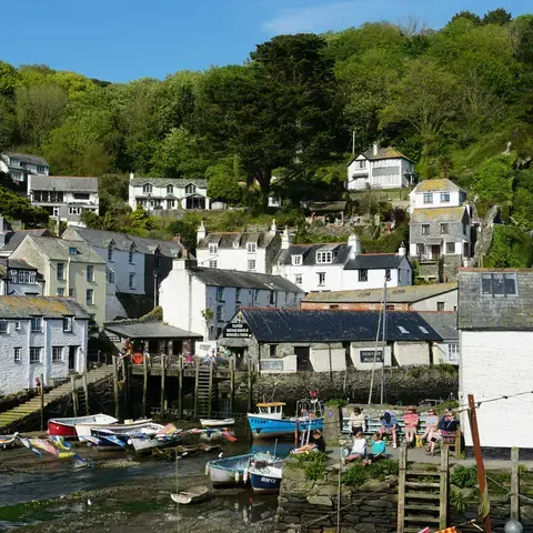 Coastal village with white cottages on a green hillside, small boats in the harbor, and people relaxing by the water