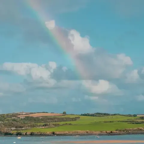 Vivid rainbow arches over lush green fields and a serene estuary dotted with small boats under a bright blue sky