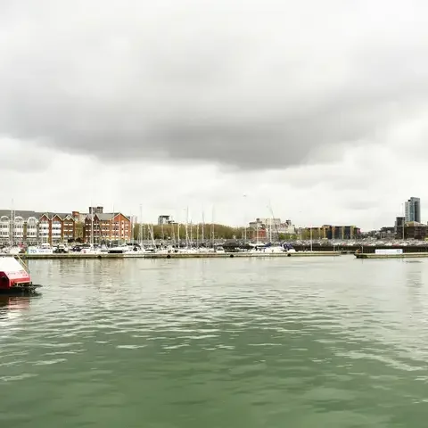 Marina with moored boats under cloudy skies, surrounded by modern and traditional buildings reflecting on calm green water