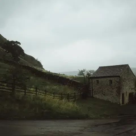 A lone stone cottage stands beside a winding road, nestled against a misty hillside with lush greenery and wooden fences