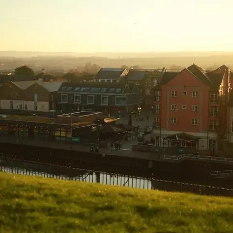 Urban riverside area with modern buildings, soft evening sunlight, and a grassy foreground overlooking the scene