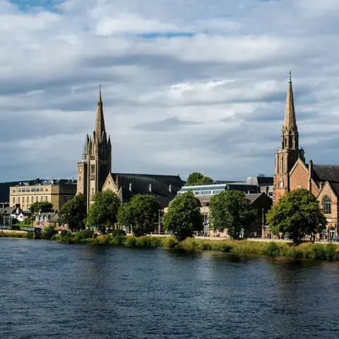 Historic riverside buildings with tall spires under cloudy skies, framed by greenery and a scenic waterfront