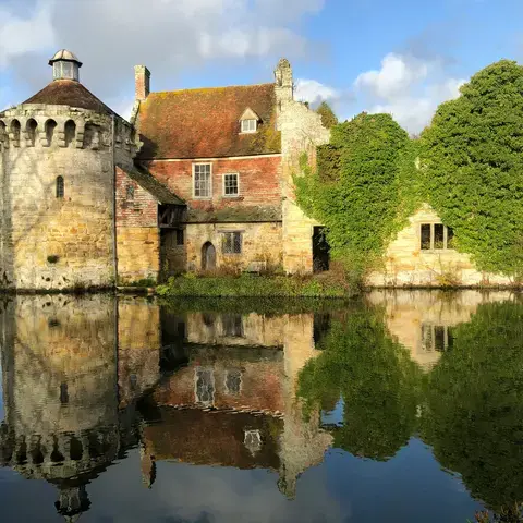 Historic stone castle with ivy-covered walls reflecting in a calm moat, under a partly cloudy sky
