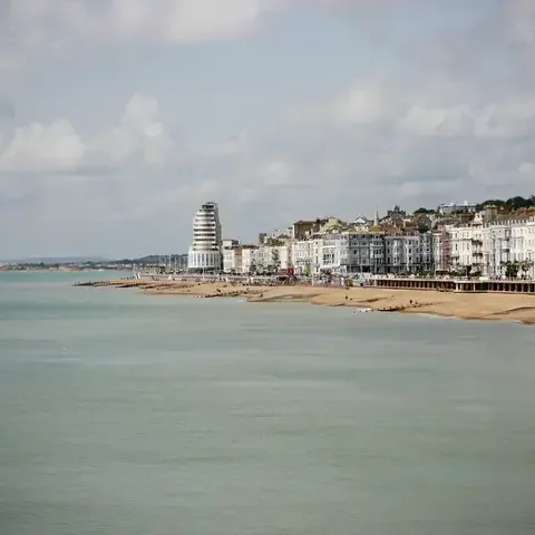 Coastal cityscape with a sandy beach, a modern cylindrical building, and Victorian townhouses under a partly cloudy sky.