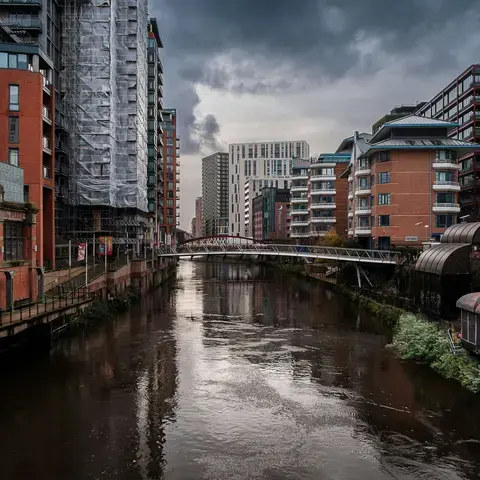 Modern buildings line the river in Manchester’s city center with a pedestrian bridge connecting both sides under a cloudy sky
