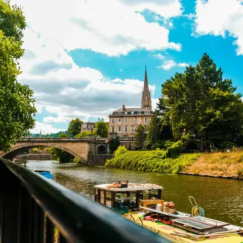 Riverside view with lush trees, a stone bridge, and a church spire rising behind historic buildings under a bright blue sky