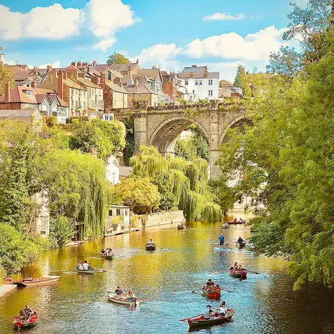 Historic stone bridge over a river surrounded by charming townhouses and lush trees, with people enjoying rowing boats