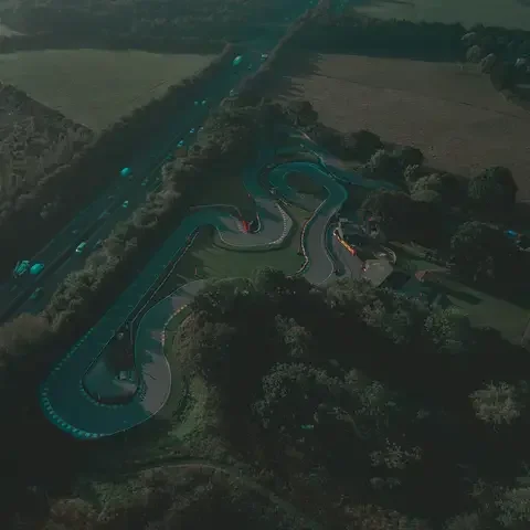 Aerial view of a winding karting track surrounded by greenery, next to a busy highway under a soft evening sky