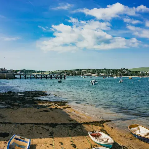 Small boats rest on a sandy shore by a harbor under a bright blue sky,with distant green hills and buildings across the water
