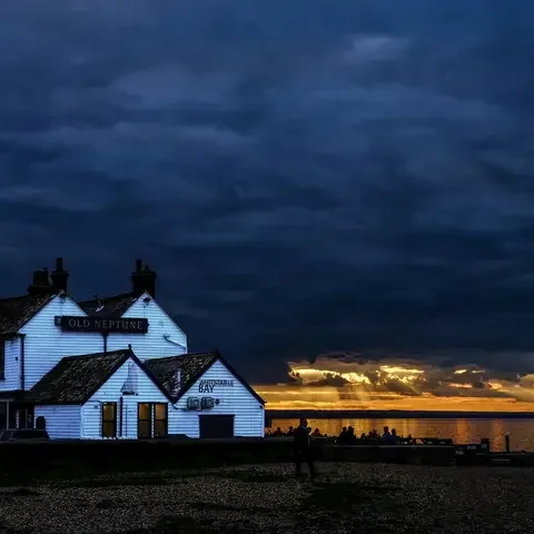 ‘Old Neptune’ pub at twilight, dramatic clouds above, golden sunset rays breaking over calm waters with silhouetted visitors.