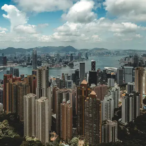Skyline of Hong Kong with its towering skyscrapers, Victoria Harbour, and lush green hills under a bright, cloudy sky