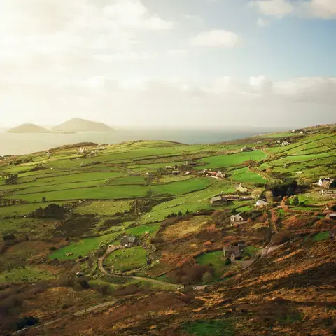 Rolling green Irish hills with scattered cottages overlooking the ocean, basking in soft sunlight under a partly cloudy sky.