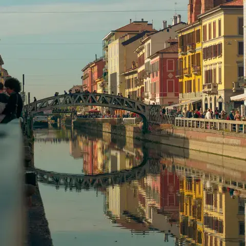 A lively Milan canal in Navigli, with colorful buildings reflecting in the water and people strolling along the promenade.