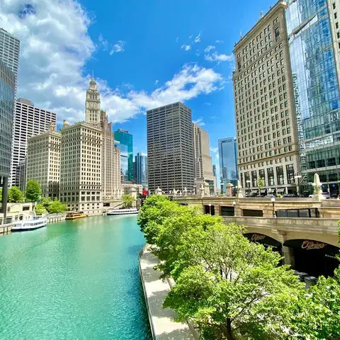Chicago Riverwalk on a sunny day, with skyscrapers reflecting on the turquoise water and lush green trees lining the riverban