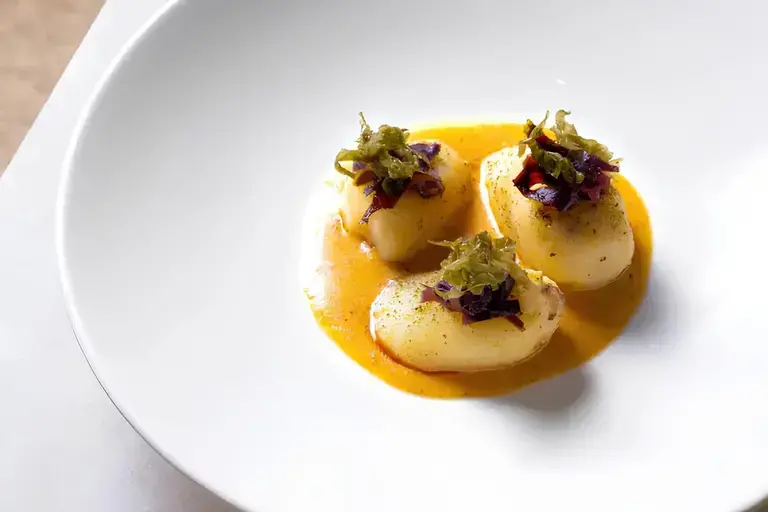 3 delicate scallops on a rich orange sauce, topped with finely chopped greens and purple garnish, arranged in a white bowl