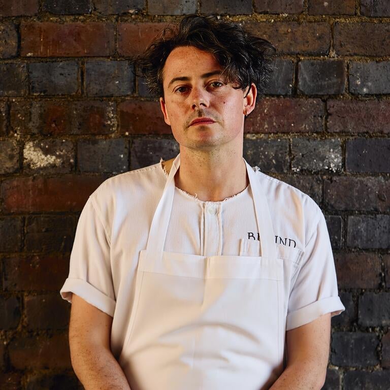 Chef Andy Beynon in a white apron, standing against a wall.