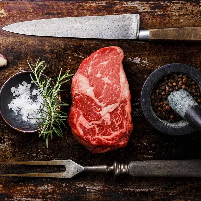 Ribeye steak on a board with seasoning, herbs, and a knife and fork.