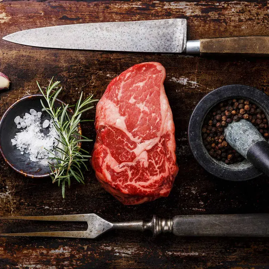 Ribeye steak on a board with seasoning, herbs, and a knife and fork.