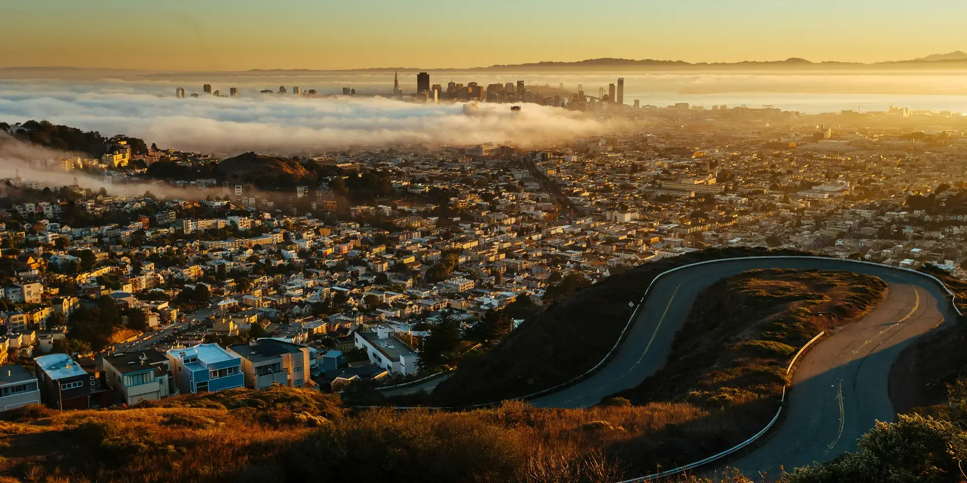 Cityscape at sunrise with fog covering the downtown area, viewed from a winding hilltop road