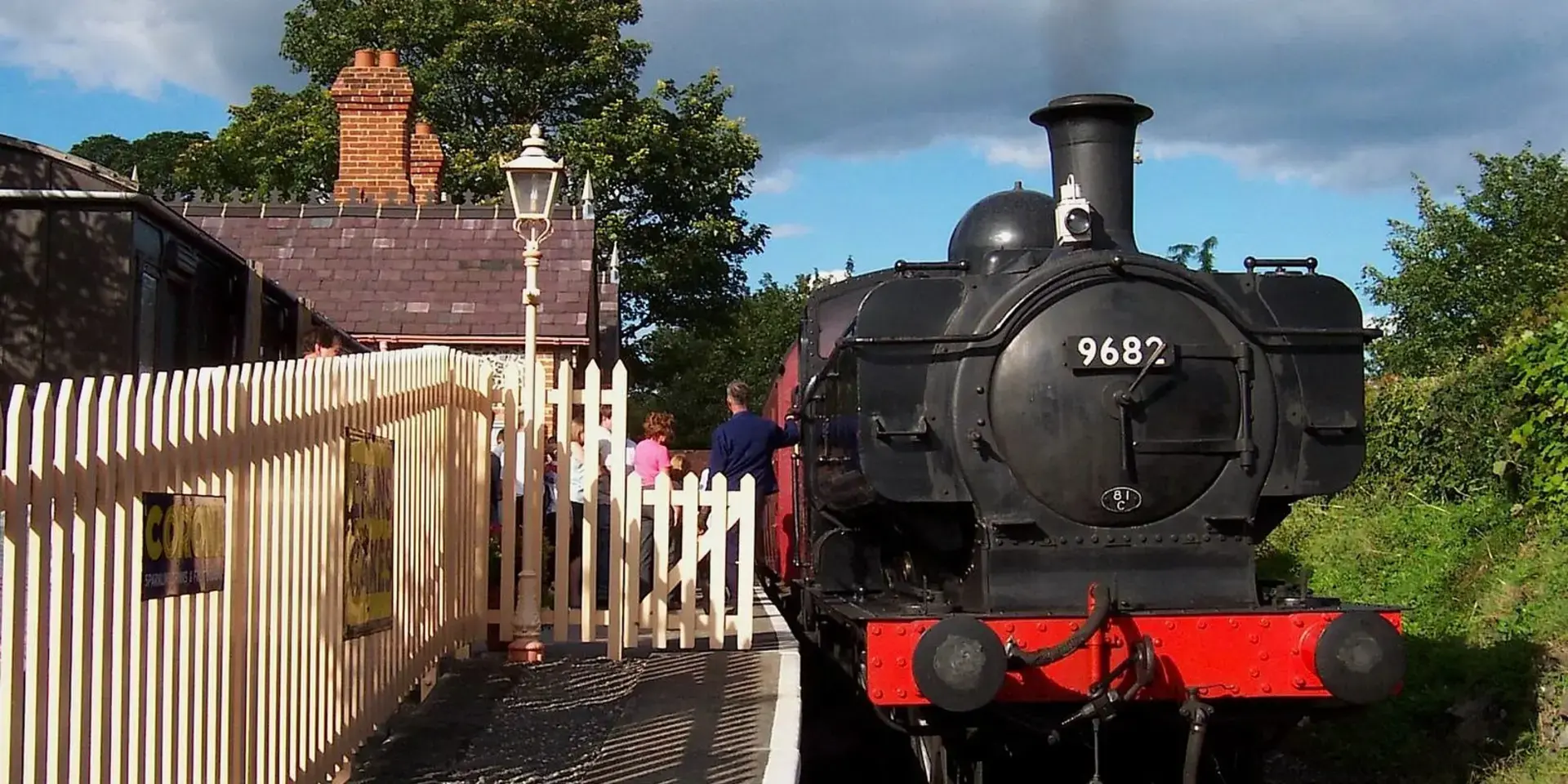 Vintage black steam locomotive number 9682, resting at a quaint station with white fences, under a partly cloudy blue sky