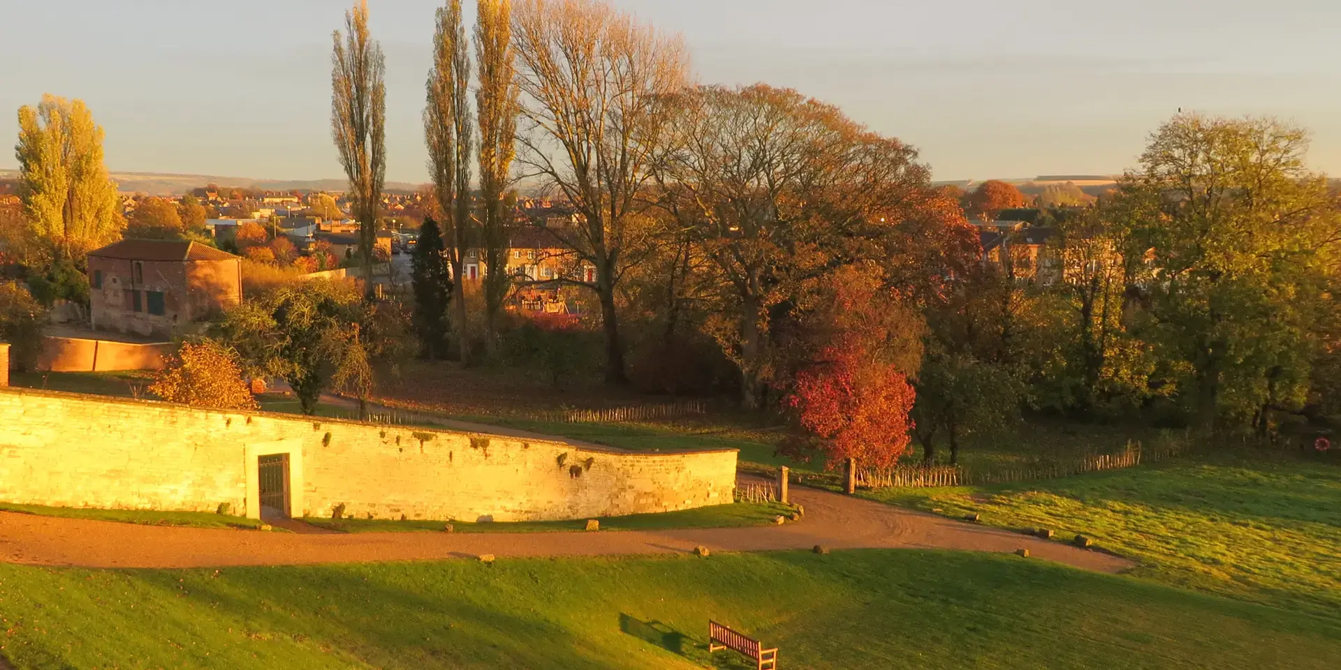 Golden hour lights up autumn trees, a stone wall, and a peaceful park bench, overlooking a quaint townscape in the distance