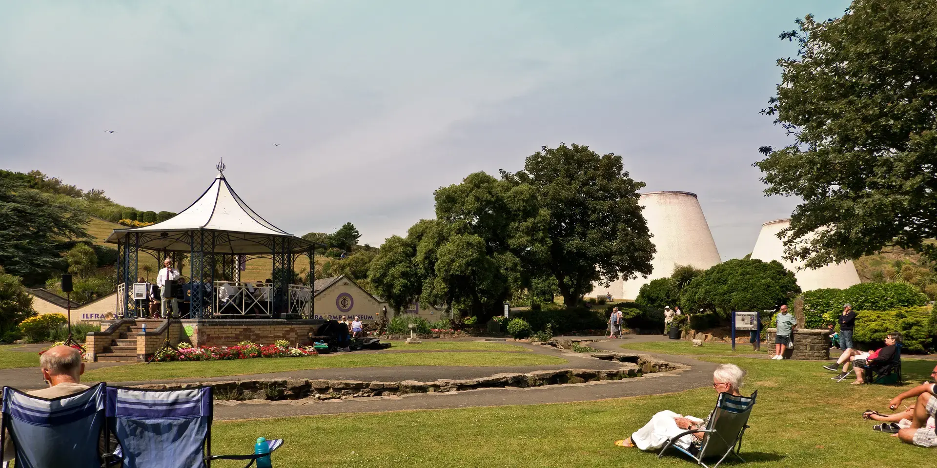 Outdoor concert in a park with a gazebo, audience seated on the grass, and trees surrounding the scene on a sunny day