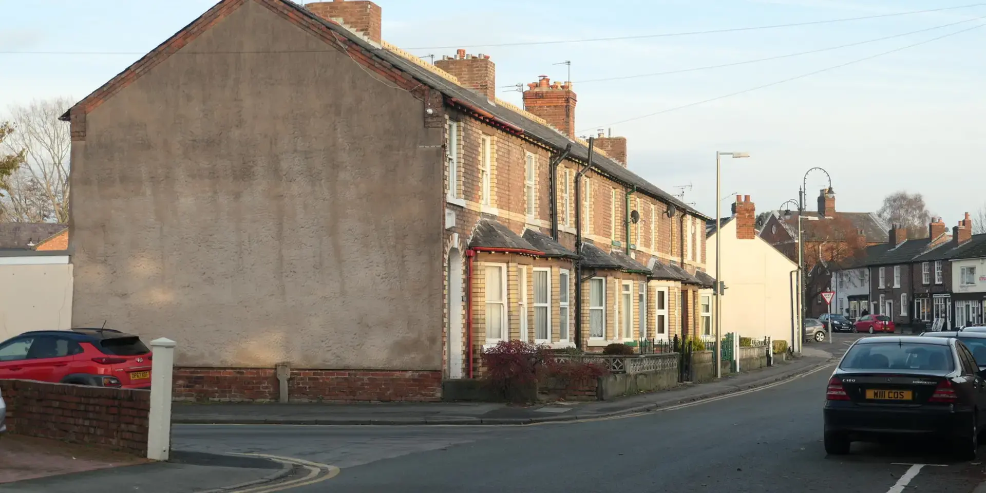 A quiet suburban street with brick terraced houses, parked cars, and a serene atmosphere under a clear sky.