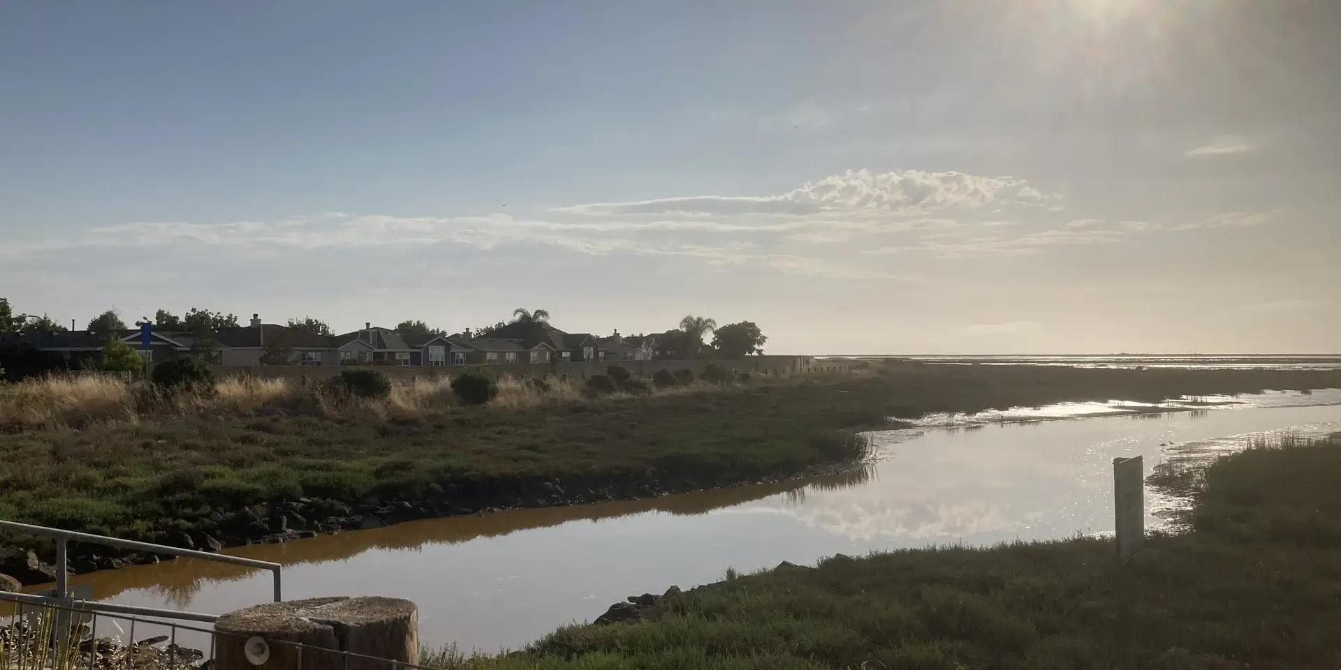 Scenic wetlands with grassy marshland, a reflective water channel, houses in the distance, and the sun shining in a clear sky