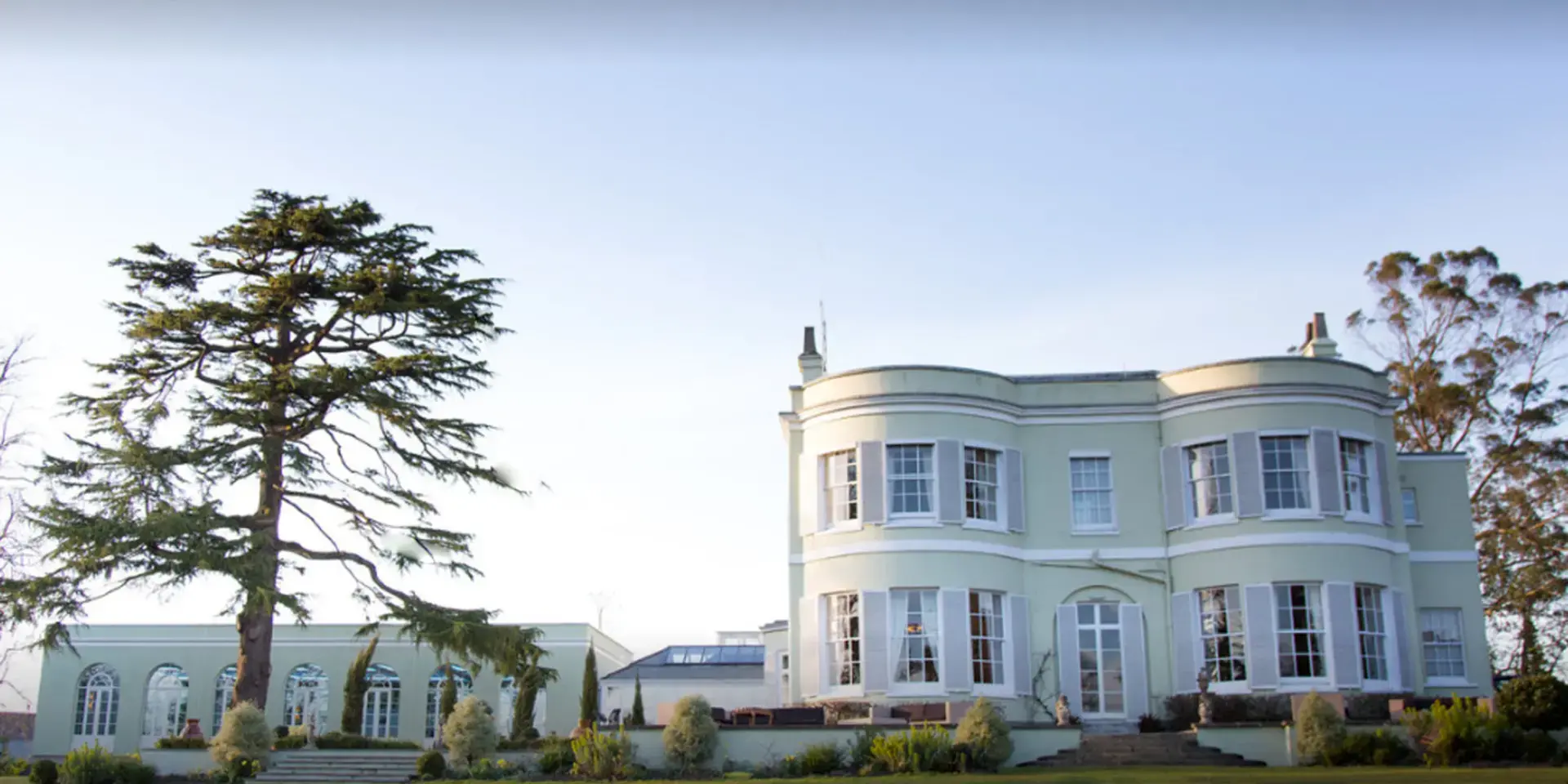 Elegant country house with pale green facade, large windows, and a garden with a tall tree under a clear sky