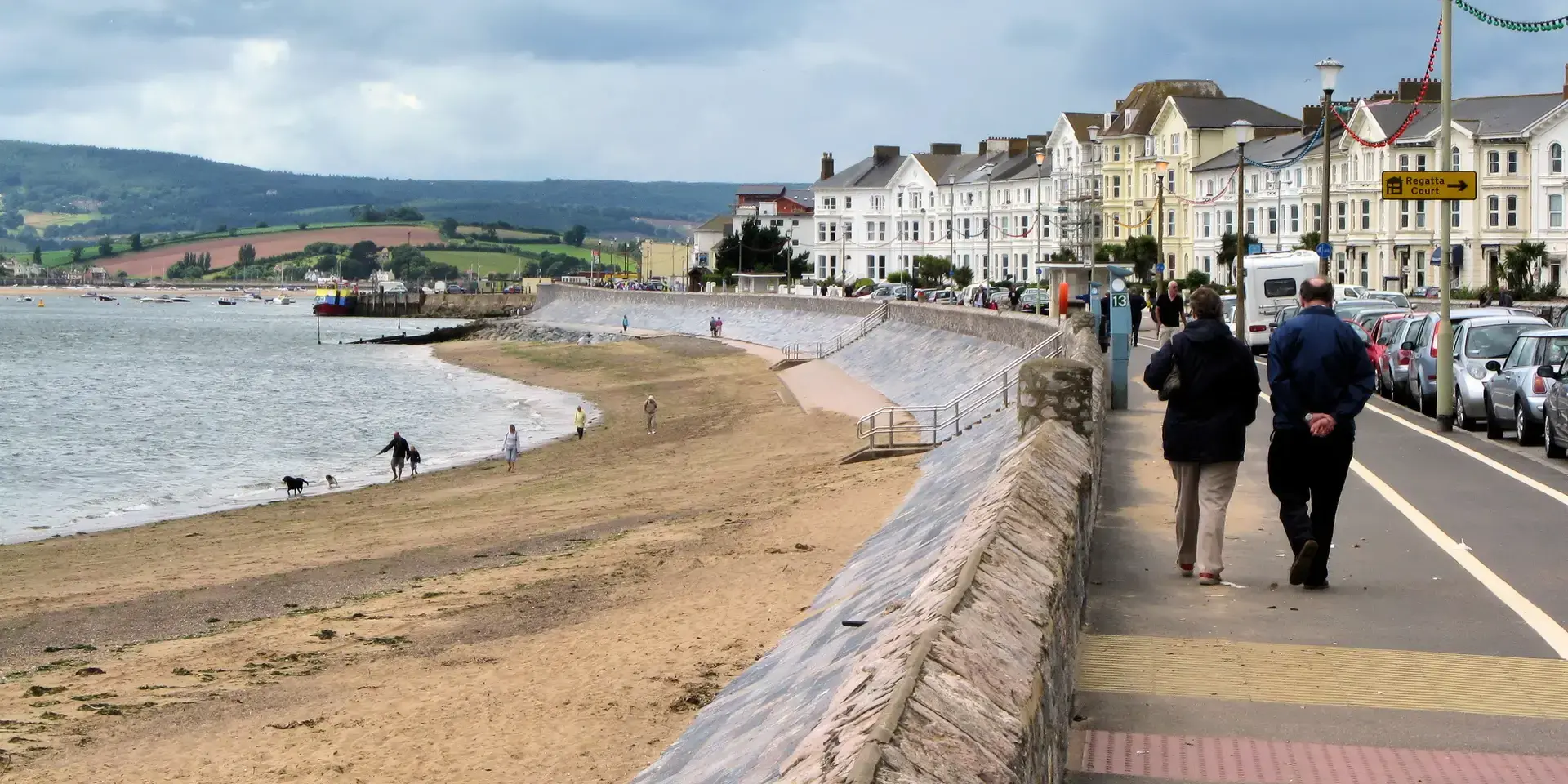 Beachfront promenade with walkers, a sandy beach, and rows of Victorian houses under a cloudy sky