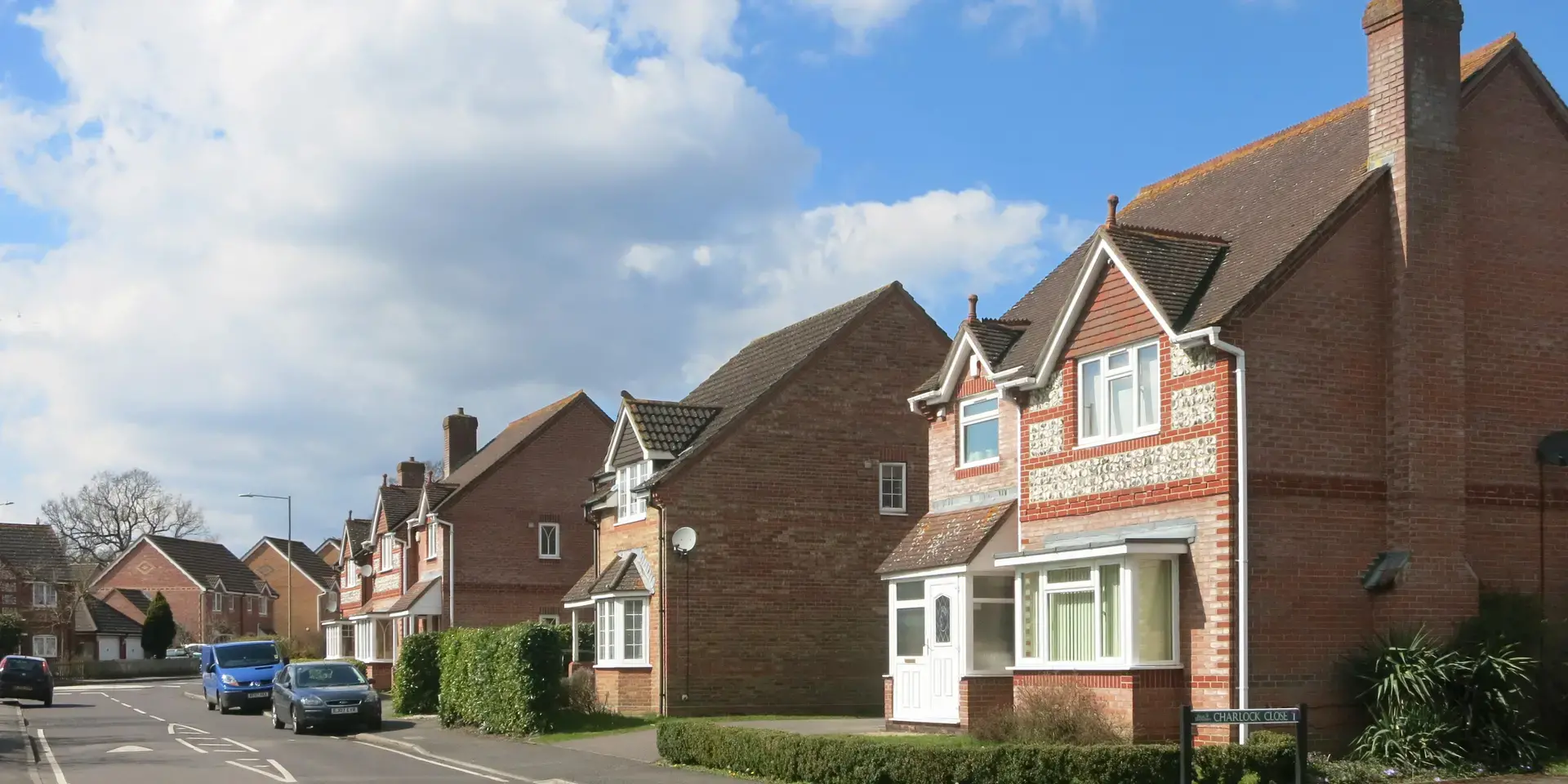 Suburban street with rows of red-brick houses, parked cars, and a partly cloudy blue sky