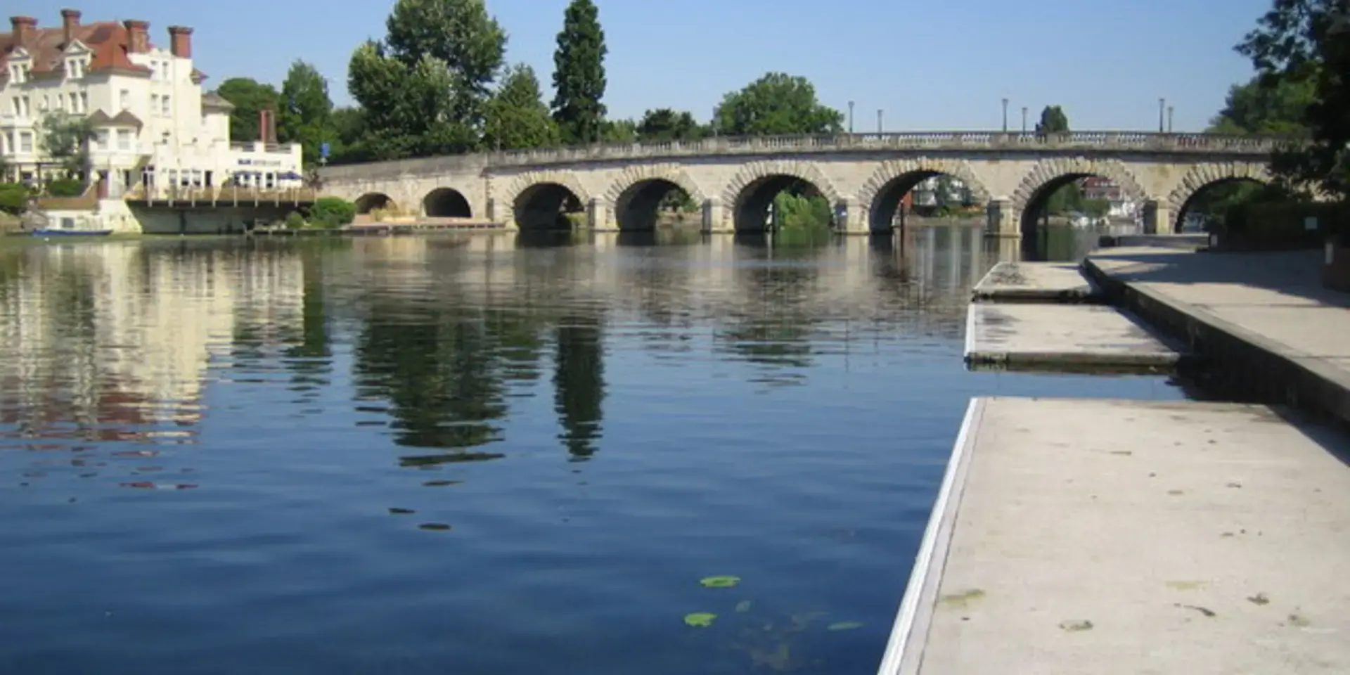 Arched stone bridge spanning a calm river with lily pads, riverside buildings, and clear blue sky