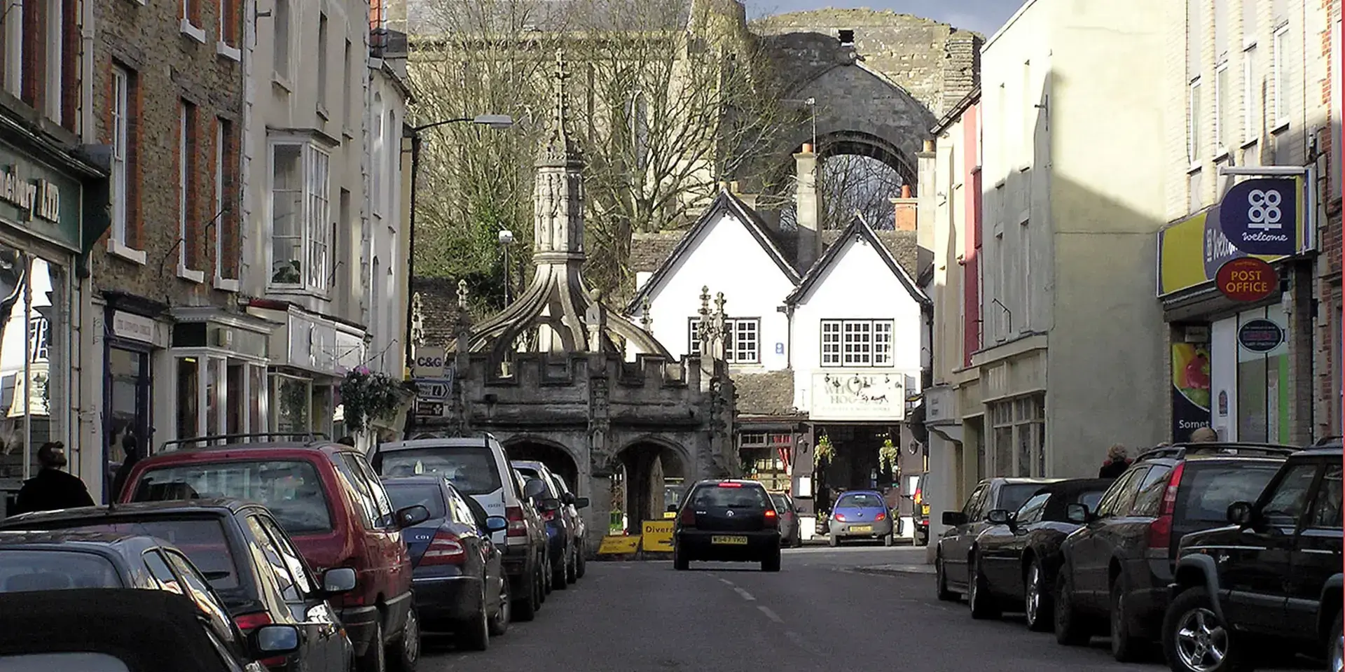 Historic town center with parked cars, medieval market cross, and ancient ruins in the background under a partly cloudy sky