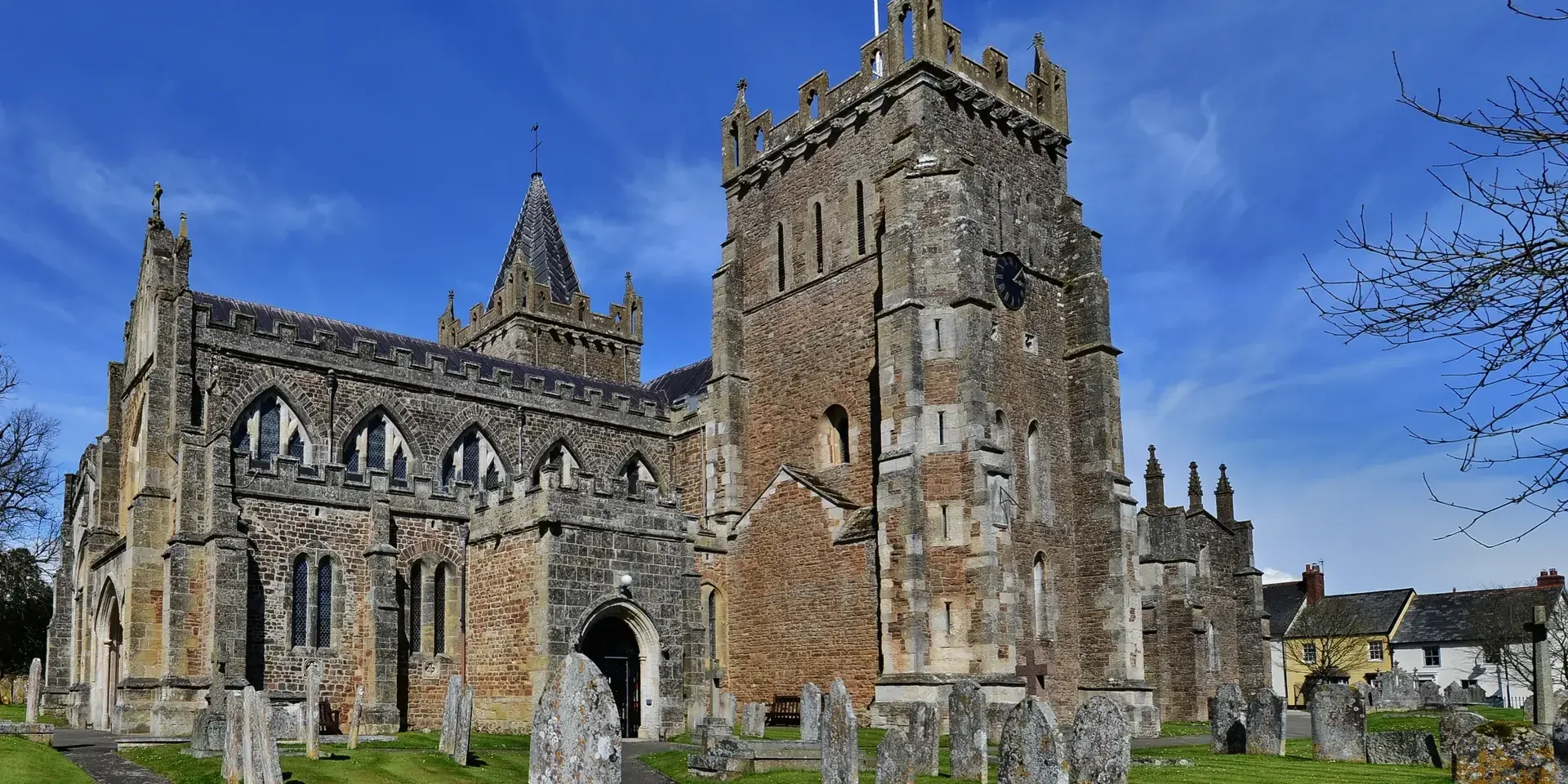 Historic parish church with Gothic architecture, a tall clock tower, and a graveyard under a bright blue sky.
