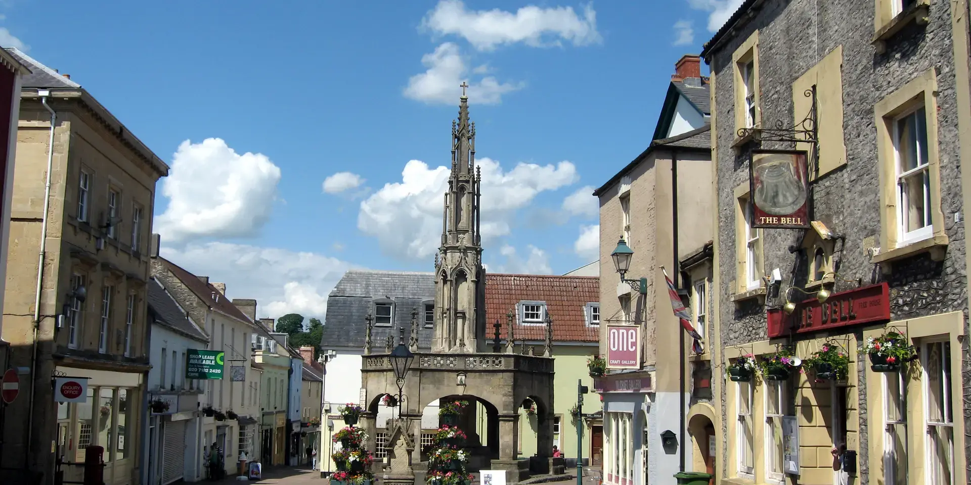 Sunny market square in a historic town with The Bell pub, shops, and a decorative stone monument under a blue sky with clouds
