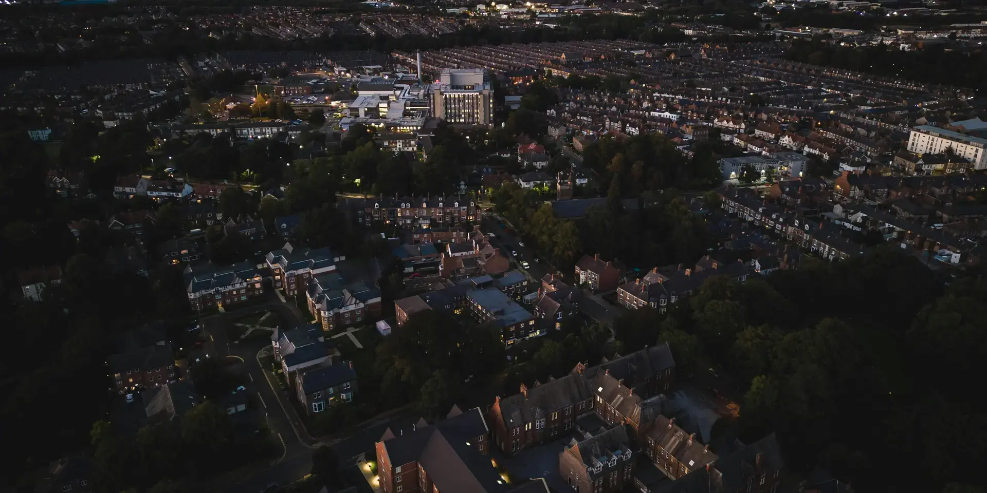 Aerial view of a quiet town at dusk, with glowing streetlights, dark rooftops, and the horizon fading into the evening sky.