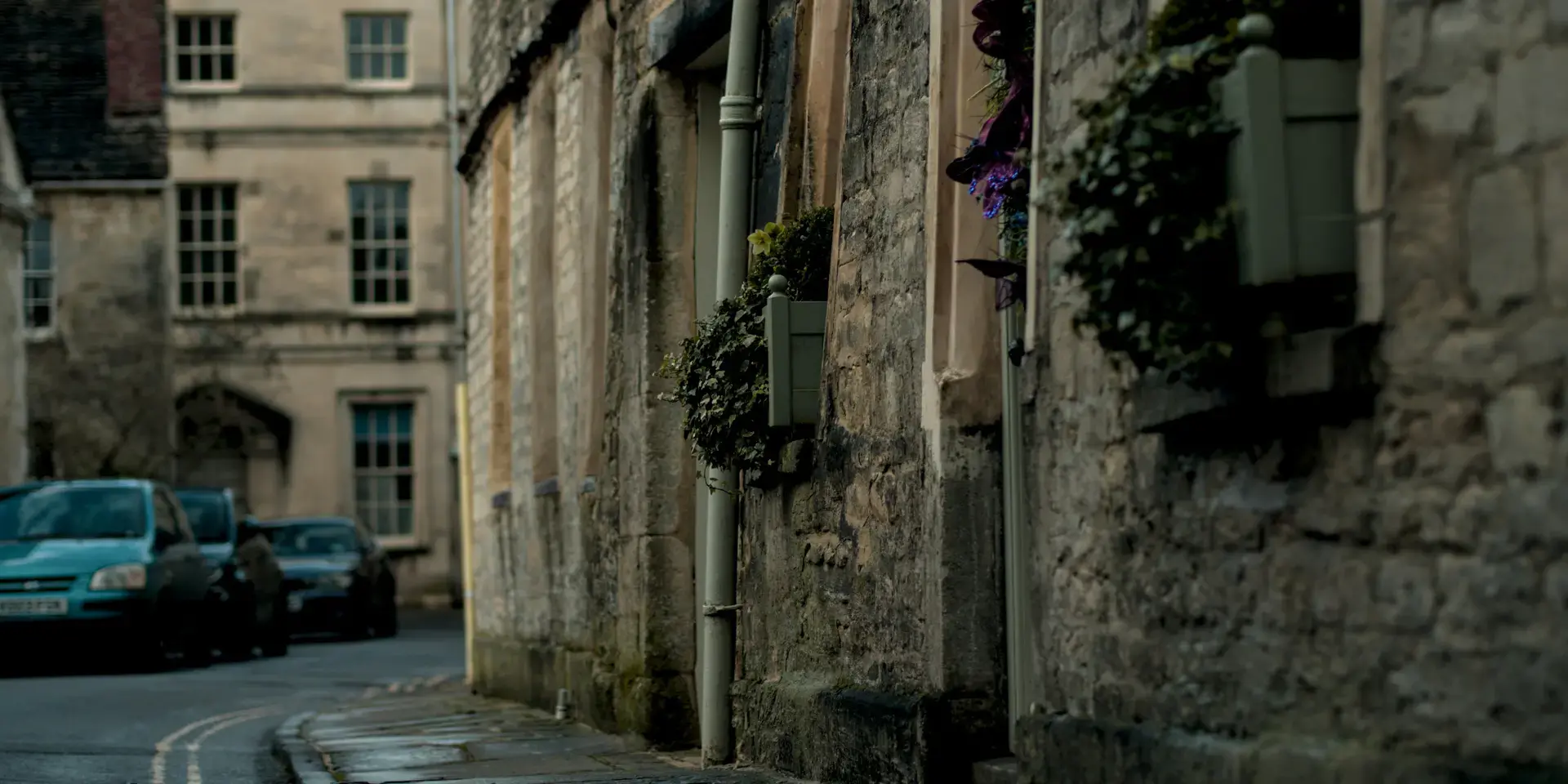 Quaint stone street with flower-filled window boxes,framed by historic architecture and parked cars under a soft,muted light