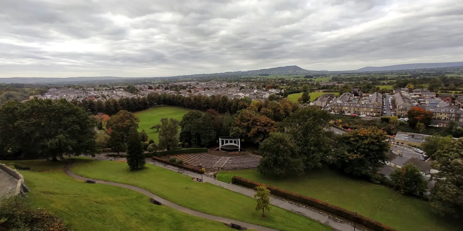 Overlooking a lush park with an amphitheater, nestled among a quiet town and rolling hills under a cloudy sky