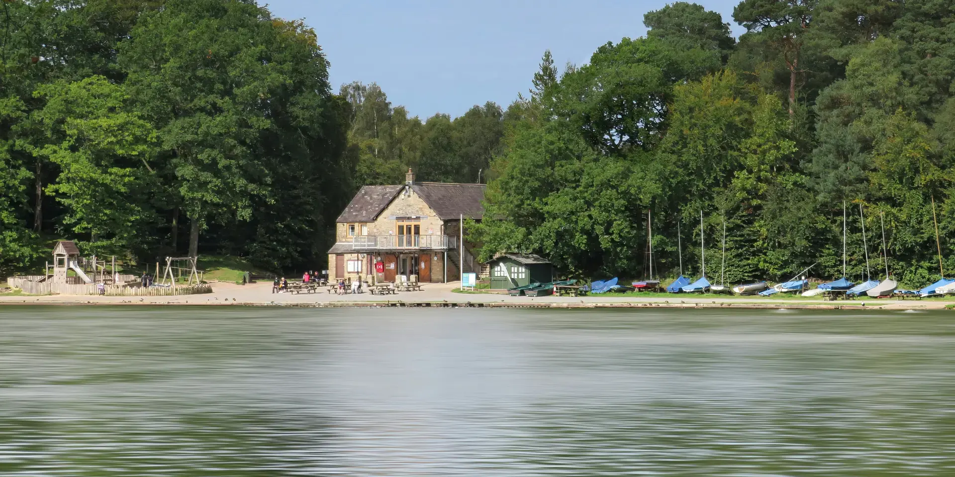 Lakefront café with a sandy beach, playground, and sailboats docked under a canopy of lush green trees on a sunny day.