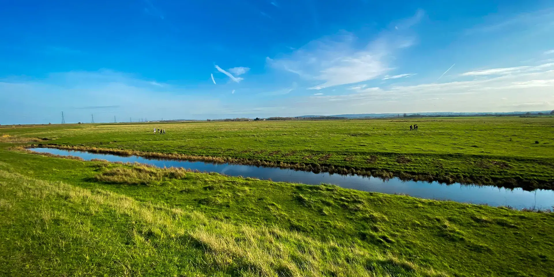 Expansive green meadow with a narrow stream under a clear blue sky, with people walking in the distance