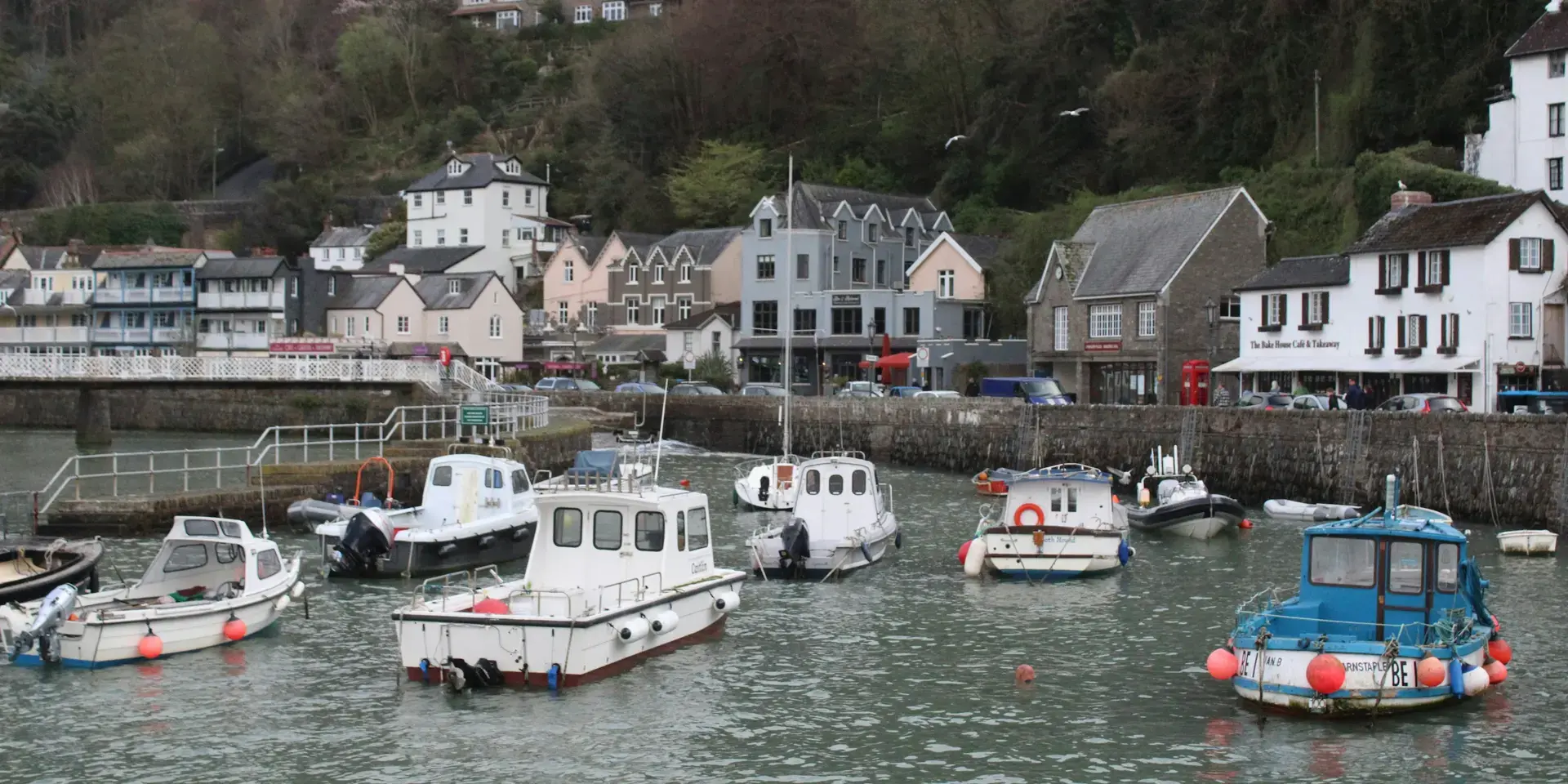Small harbor with fishing boats, colorful houses, and lush hills in the background on a cloudy day.
