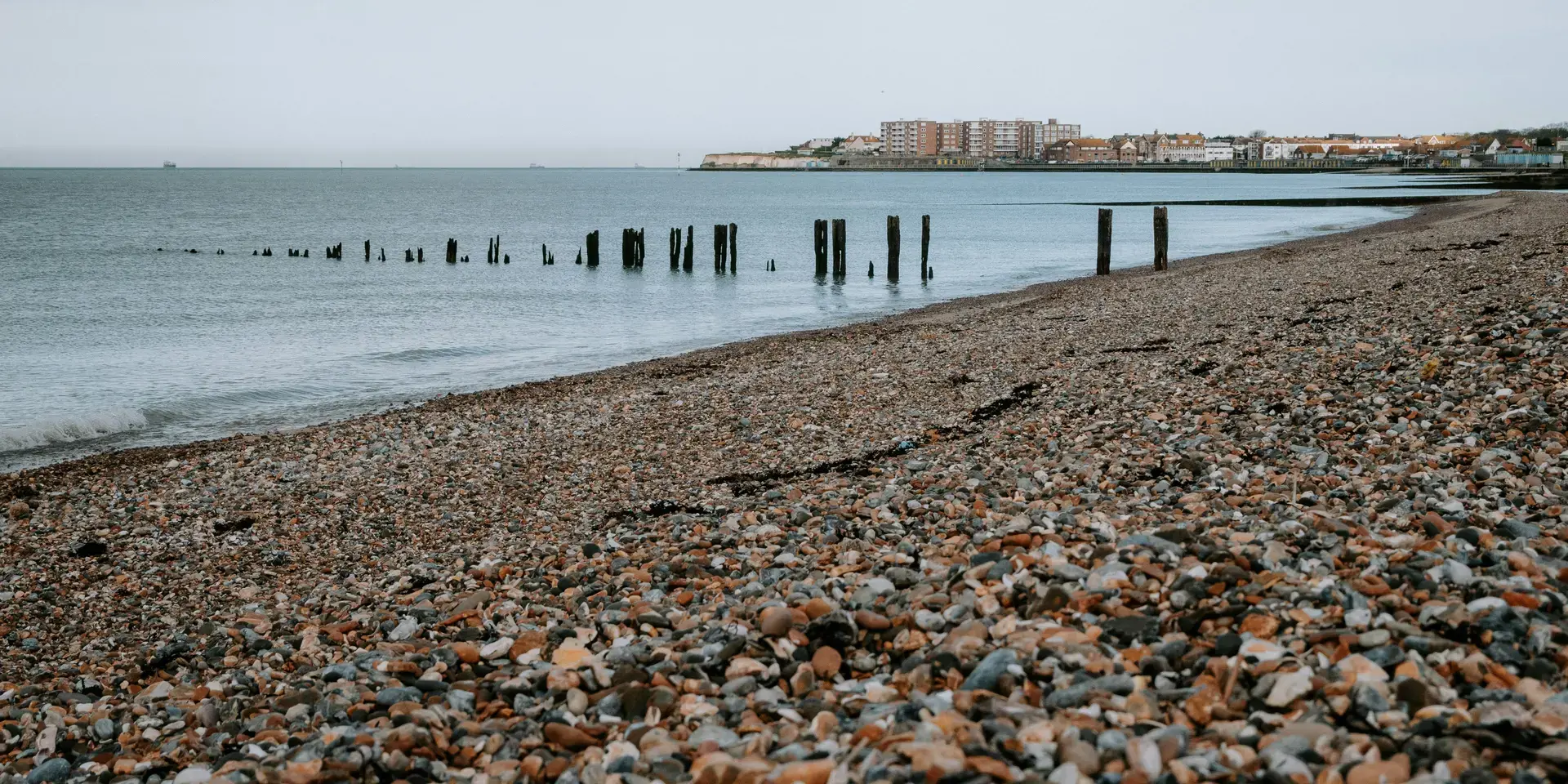 Pebble beach with weathered wooden posts in the water, overlooking distant buildings along the shoreline