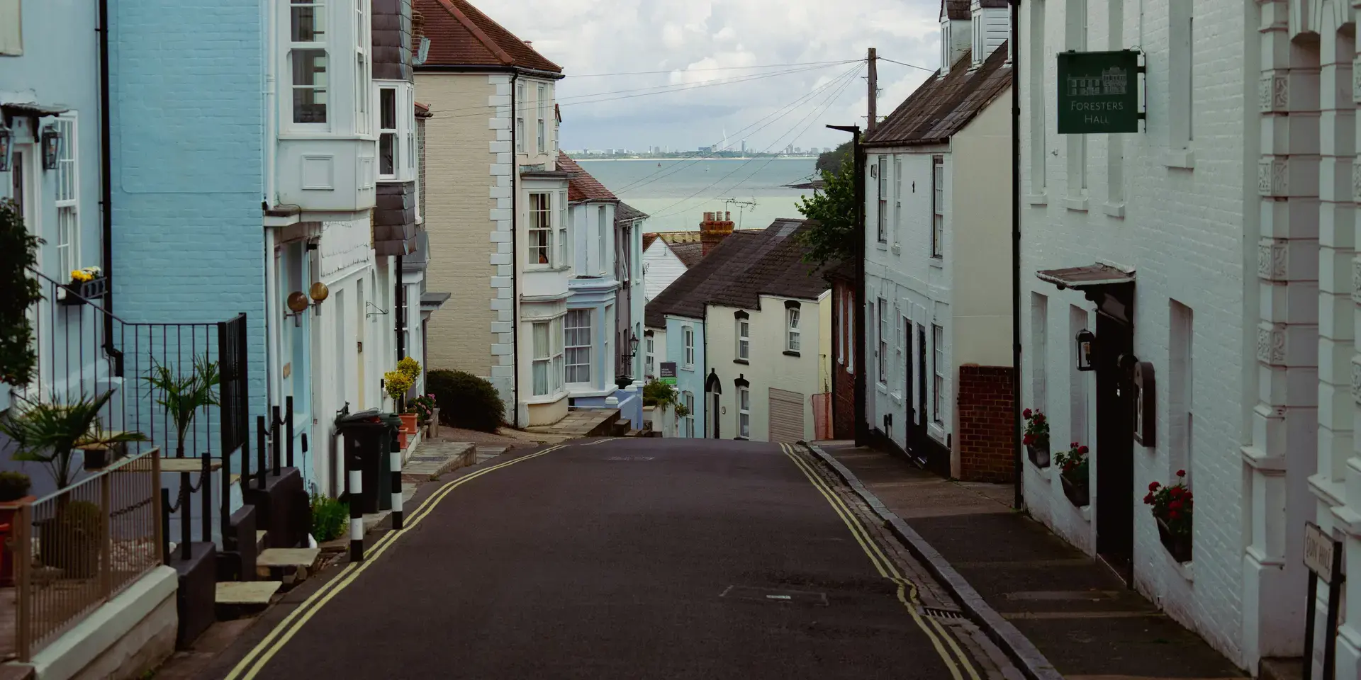 A narrow coastal street lined with pastel-colored houses leading to a serene view of the sea under a cloudy sky.