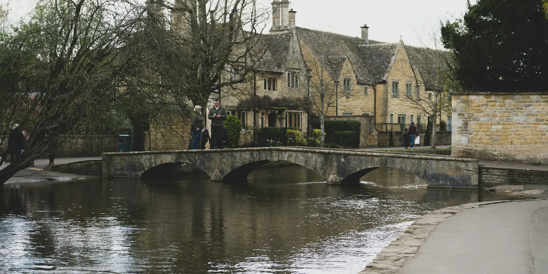 A stone bridge arches over a tranquil stream in a quaint village, flanked by charming cottages and budding spring trees