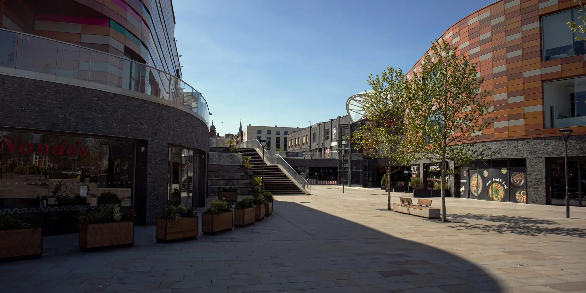 Modern city square with colorful architecture, glass-fronted buildings, stairs, trees, and restaurants under a clear blue sky